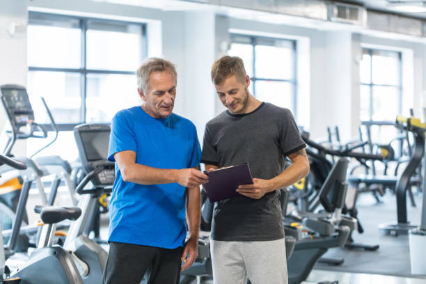 Fitness-Plan-001 Male trainer showing fitness progress report to senior man at rehab, both looking at clipboard. Old man discussing rehabilitation program with instructor at gym.