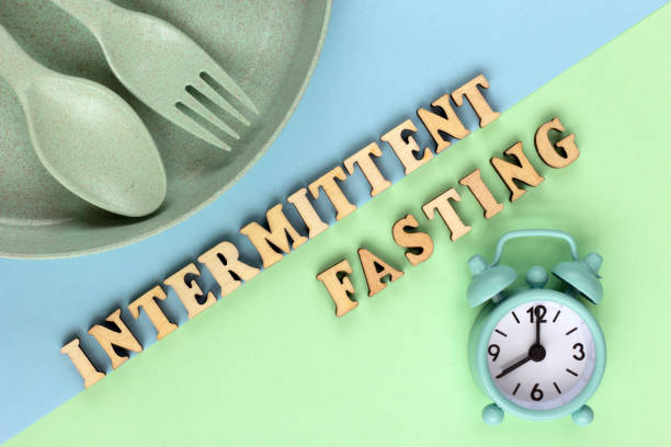 Intermittent-Fasting-001 Intermittent, keto, detox diets and weight loss concept: clock, plate, spoon and fork. The word Intermittent fasting from wooden letters. The concept of proper nutrition.