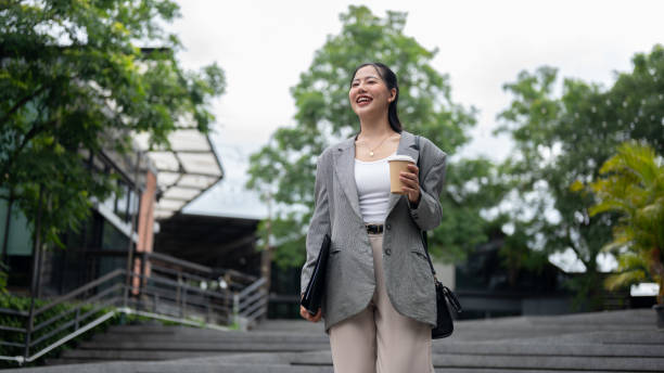 A beautiful, confident Asian businesswoman in a stylish suit is walking down the stairs in the city with a takeaway coffee cup in her hand, going to the office in the morning.
