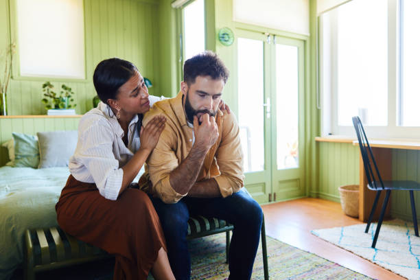 Young woman sitting with her arm around her upset husband while consoling him on their bed at home