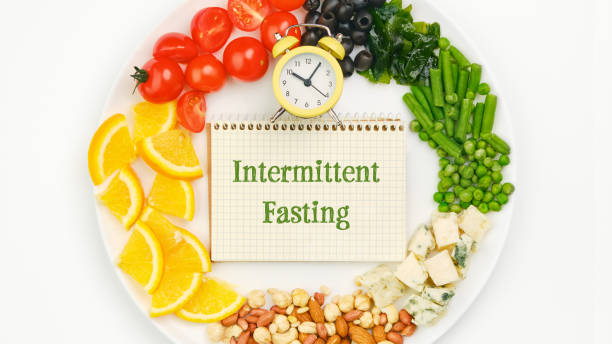 Circle of assorted fresh food on a plate. Clock and notebook with the text Intermittent Fasting. Diet, weight loss and lunchtime concept.