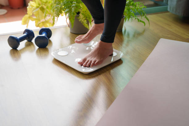 A close-up view of a young woman standing on a weighing scale, focusing on her feet and the display as she checks her weight. The scene highlights the importance of health and self-awareness in maintaining a balanced lifestyle.