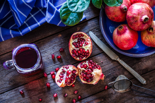 Top view of a rustic wooden table with a glass jar filled with pomegranate juice. Some pomegranates are in a blue plate and one is cut in half. A blue napkin is at the top-left corner. Some seeds are spilled on the table.