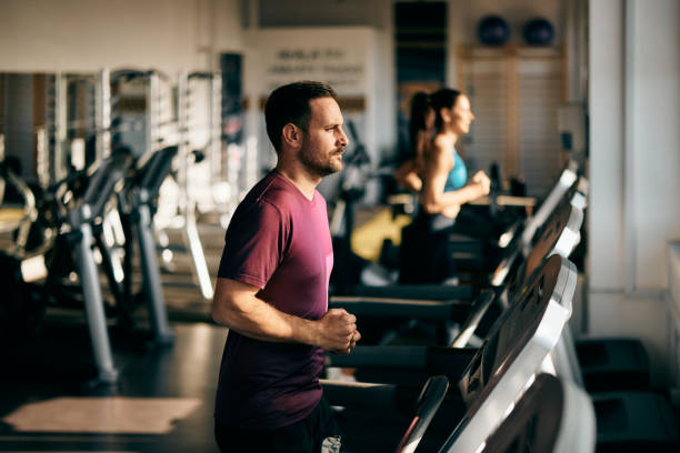 Side-view of a male runner, running on a treadmill, at the gym, a girl running in the background.
