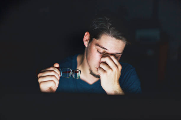 A young man, seen at night against a black background has taken off his glasses and is rubbing his eyes. He could be very tired or having problems with sore eyes.
